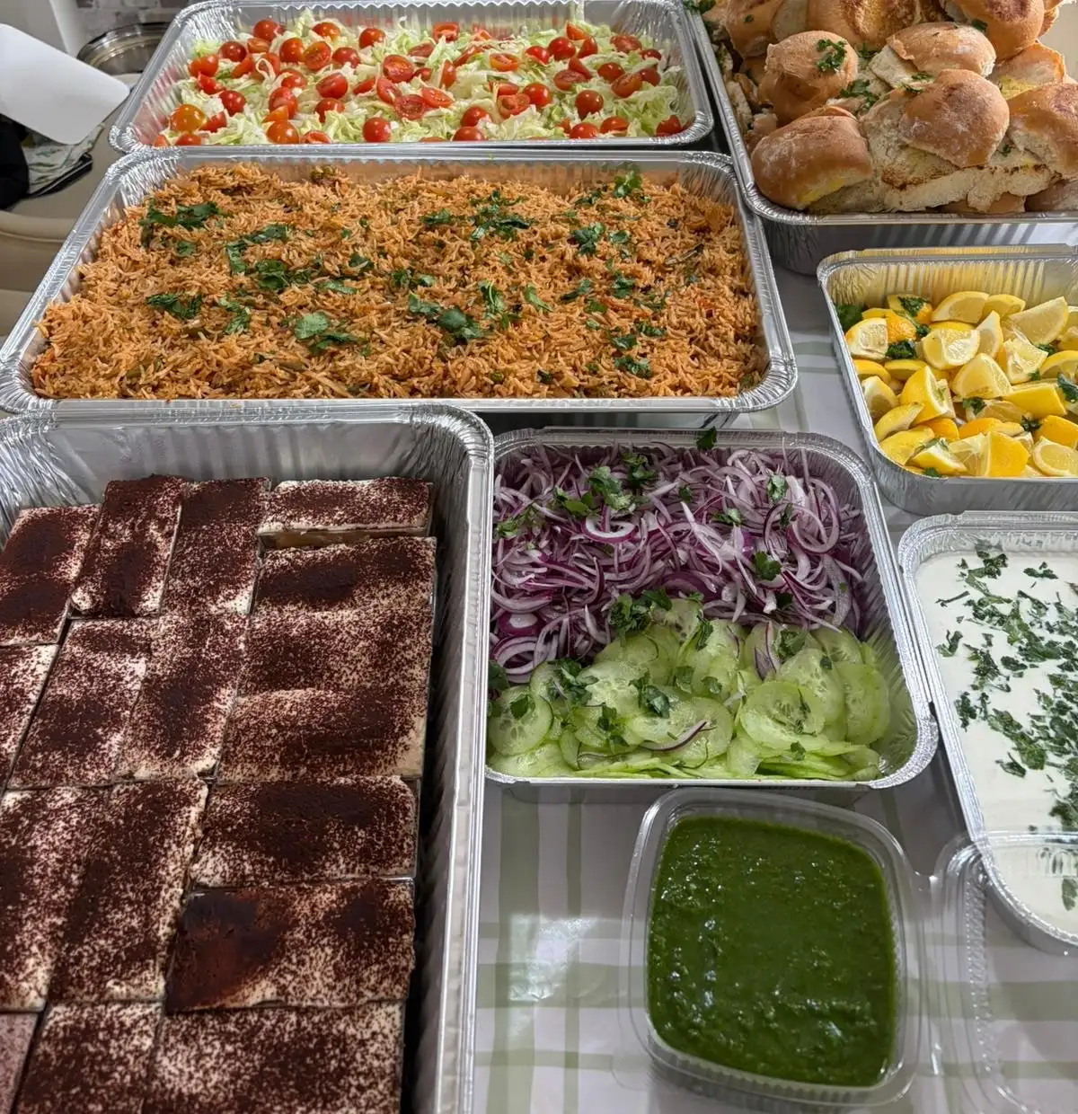 A catering spread in foil trays: dal makhani, a tomato and cabbage salad, tawa pulav, toasted pav, lemon wedges, red onion and cucumber, green chutney, raita and tiramisu.