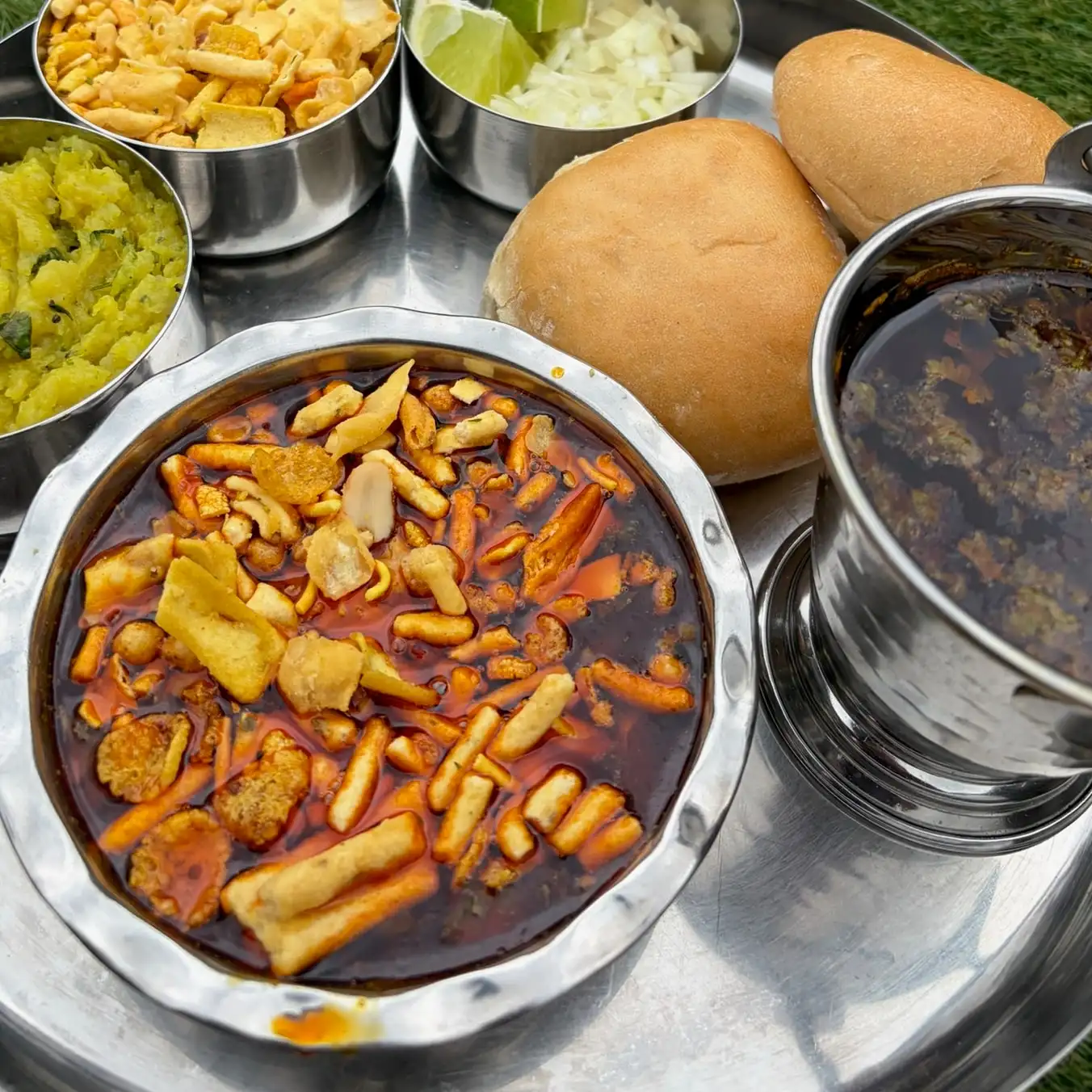 A steel thali of missal pav from above: missal, farsan, pav, onion and lime, potato bhaji and a cup of tarri.