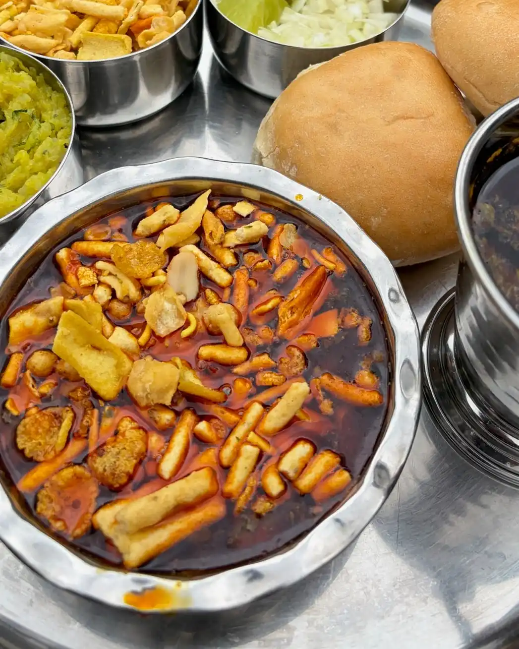 Missal pav on a steel thali: a bowl of red sprouted-bean curry topped with farsan, with pav, onion, lime and potato bhaji.