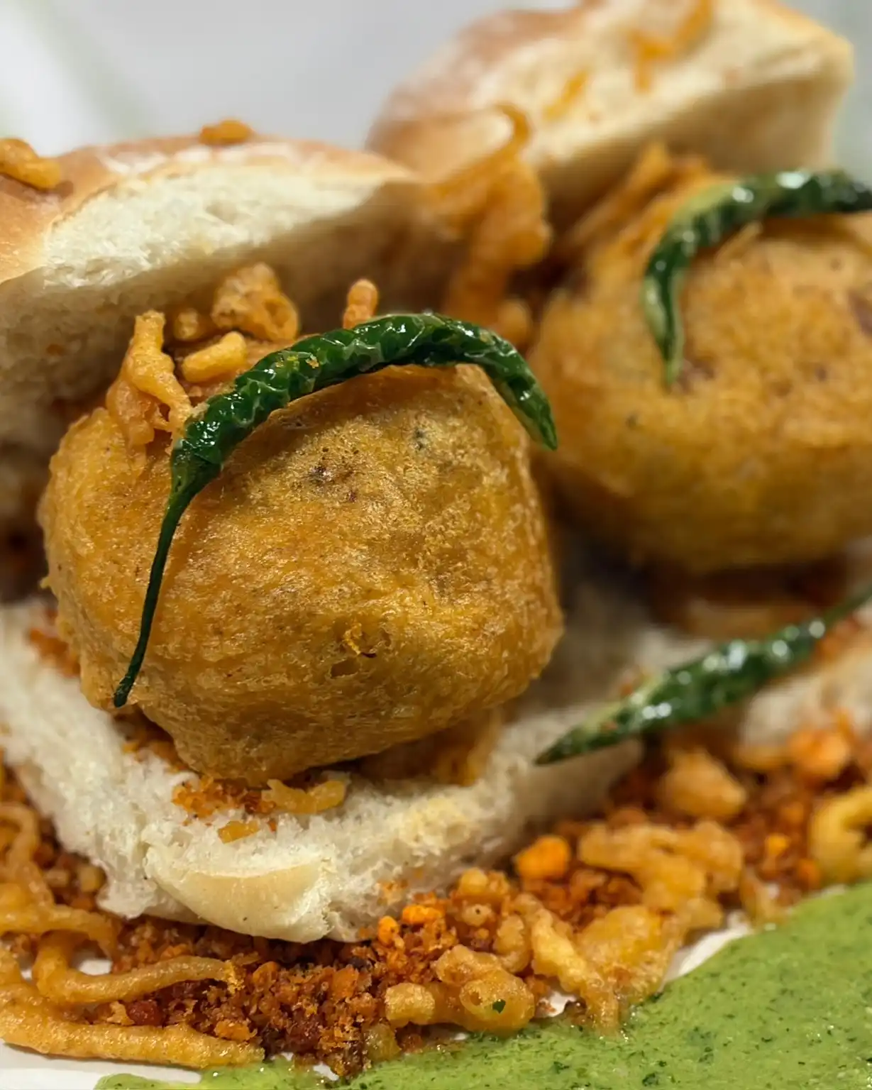 Two vada pav on a white plate, each with a fried green chilli, on dry garlic chutney with a swirl of green chutney.